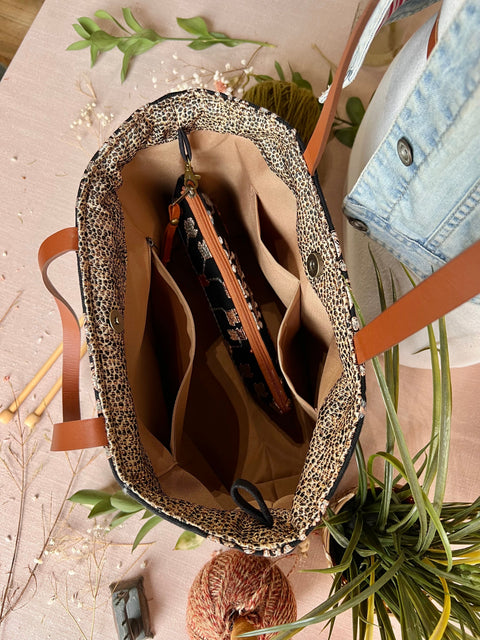 Open tote bag , beige interior, multiple compartments, and a black and orange zippered pouch inside, on a pink surface with green plants and dried flowers around.