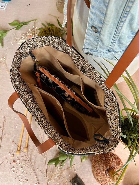 Inside of tote bag with tan leather handles, open to reveal beige interior with multiple compartments, on a pink surface with dried flowers and greenery