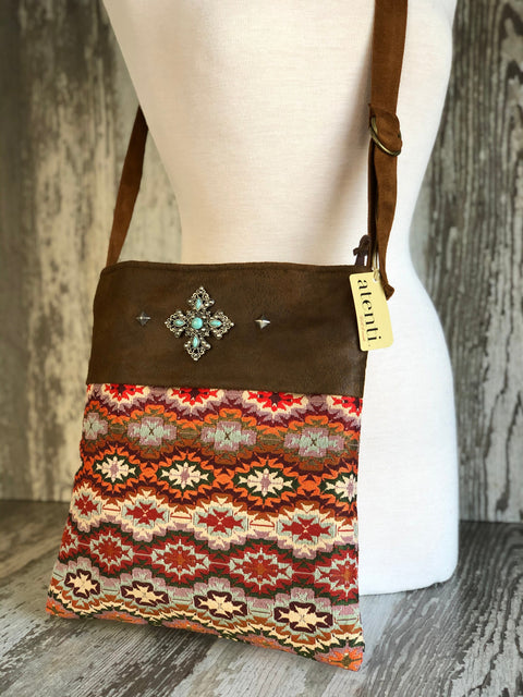 A brown shoulder bag with a geometric patterned lower half, displayed on a white mannequin torso. The bag features a brown suede-like top panel with a jeweled cross motif, a hanging tag reading 'saintelli', and a brown strap.