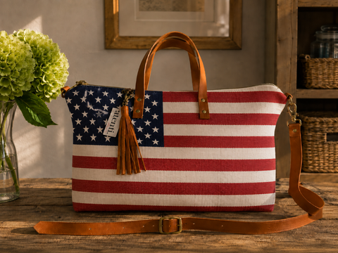 An American flag themed canvas shoulder bag with tan leather handles and strap, resting on a rustic wooden table with a vase of green hydrangeas in the background.